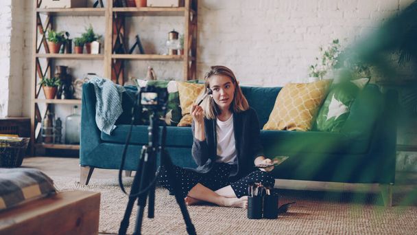 Popular female blogger is recording video about make-up and beauty sitting on floor at home and using camera on tripod. Girl is showing professional cosmetics kit and using brush. - Foto, Imagen