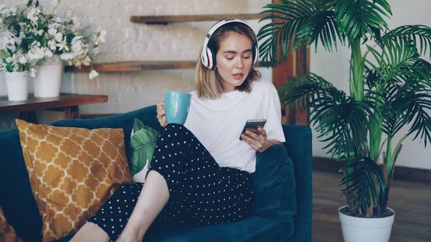 Modern young woman is resting on comfortable sofa at home with smartphone, cup of tea and headphones. Cute girl is listening to music, singing and enjoying drink. - Photo, image
