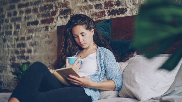 Cheerful girl is reading funny book in bedroom lying on comfy double bed, smiling and holding cup with drink. Hobby, happy people, beautiful interiors and literature concept. - Foto, Imagem