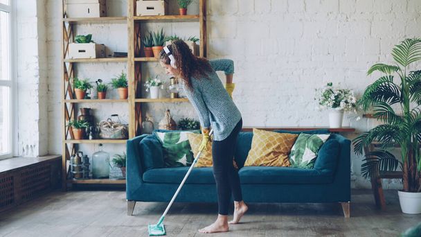 Pretty girl with dark curly hair wearing casual clothing is mopping floor at home and listening to music through headphones. Clean-up, modern houses and people concept. - Foto, immagini