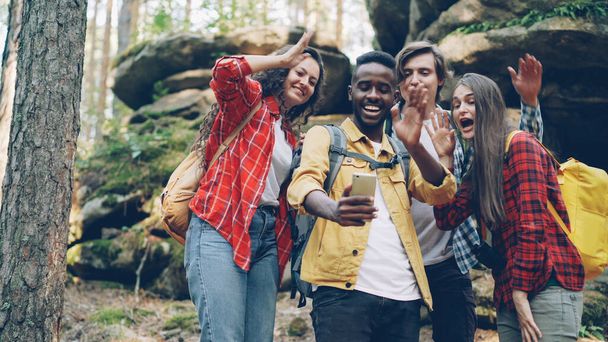 Chico afroamericano feliz está haciendo videollamadas usando un teléfono inteligente moderno durante la caminata en el bosque con amigos, los jóvenes están mirando la pantalla, saludando con la mano y hablando
. - Foto, imagen