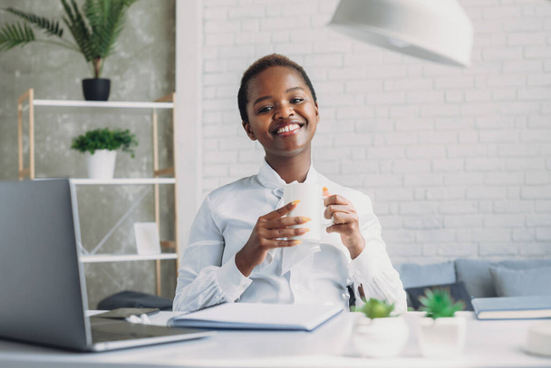 Carefree inspired african-american businesswoman sitting in modern drinking coffee, looking at the camera with friendly smile. Employee freelance online - Photo, Image