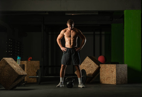 Full-length photo of a handsome man with a naked torso exercising with a kettlebell on a dark background - Fotografie, Obrázek