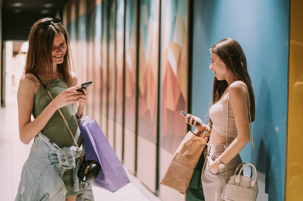 Zwei schöne beste Freundinnen halten ihre Telefone in der Hand, während sie im Flur des Einkaufszentrums stehen - Foto, Bild
