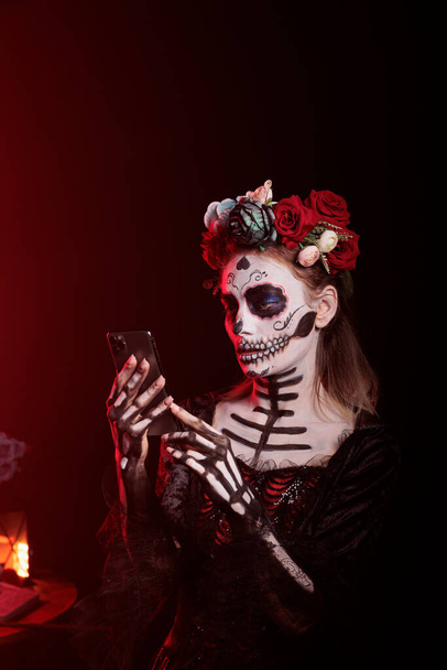黒の背景にスマートフォンを使い、スタジオでインターネットサイトを閲覧する美しい不気味な女性。メキシコの休日のお祝いに死の恐怖の女神を描いたサンタモデル. - 写真・画像