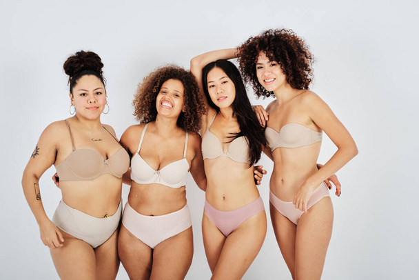 Modelos femeninos multirraciales positivos en lencería mirando a la cámara mientras están de pie juntos contra el fondo gris en el estudio - Foto, imagen
