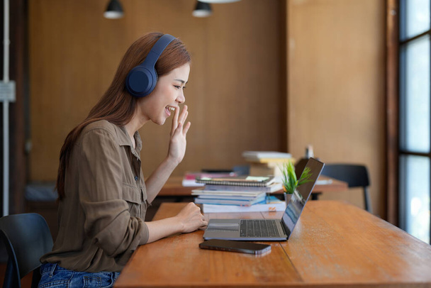 Asian tutor of language start online lesson with trainee wave hand smiling looks at pc screen. woman in headphones communicating distantly, e-learning process, application usage concept. - Photo, Image