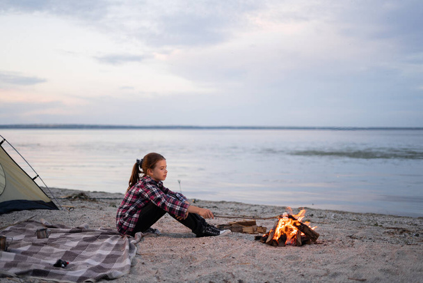 Маленькая девочка смотрит на костер, сидя на пляже в осенний вечер, небо и вода фон, ребенок в природе - Фото, изображение