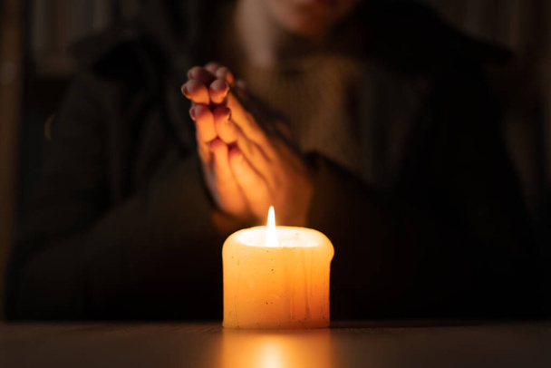 Close up womans hands dressed in a warm winter jacket sit at home at a table and warms his hands from a burning candle. Concept of no heating and electricity in winter at home. - Foto, Imagen