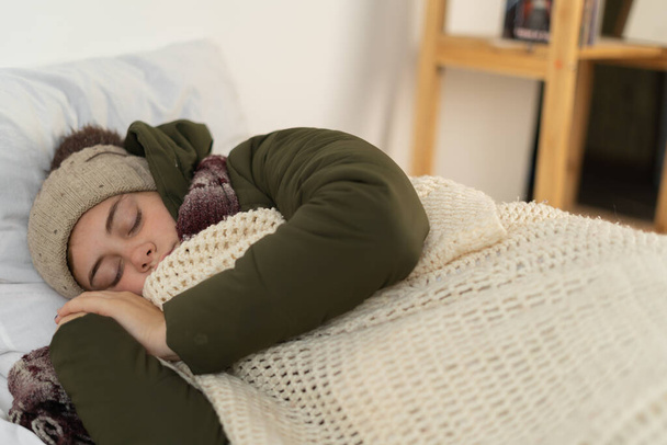 Νεαρή γυναίκα που κοιμάται με ζεστά ρούχα καλυμμένη με κουβέρτα. Κρύο χειμώνα. Δεν υπάρχει θέρμανση στο σπίτι. Χωρίς έννοια θέρμανσης. - Φωτογραφία, εικόνα