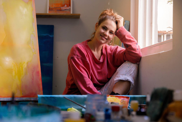 Portret białej malarki siedzącej przy oknie swojego studia patrzącej w kamerę - Zdjęcie, obraz