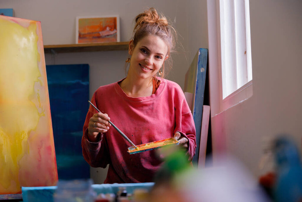 Porträt einer kaukasischen Malerin, die am Fenster ihres Ateliers sitzt und in die Kamera blickt - Foto, Bild