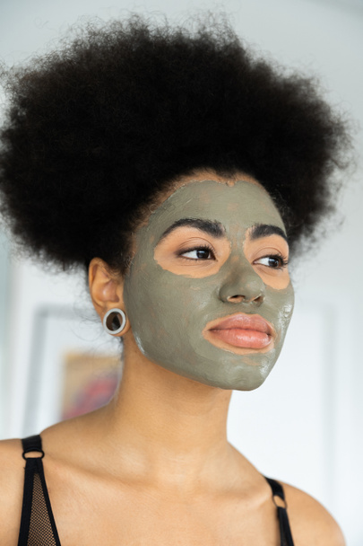 mujer afroamericana rizada con máscara de arcilla en la cara mirando hacia otro lado en casa - Foto, imagen