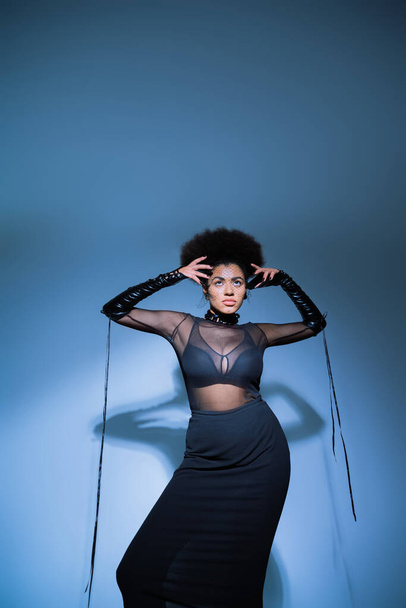 stylish african american woman in transparent shirt and black bra looking up on blue - Photo, Image