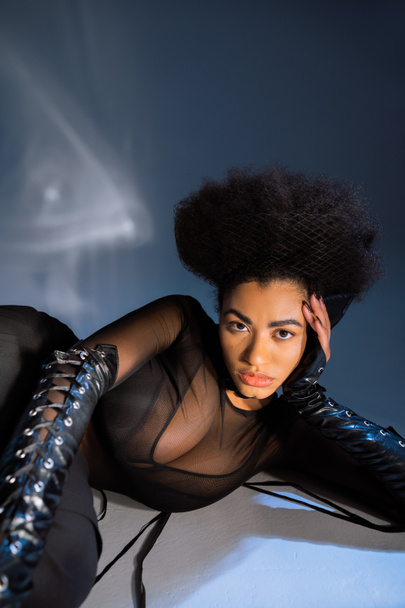 curly african american woman in transparent shirt and black bra lying on blue  - Foto, imagen