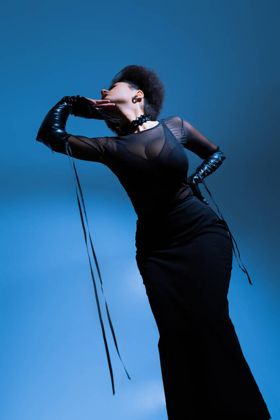 low angle view of curly african american model in black outfit posing with hand on hip on blue - Fotografie, Obrázek