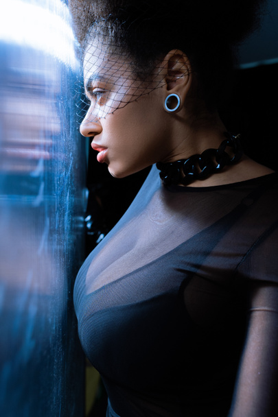 junge afrikanisch-amerikanische Frau in sexy Outfit und schwarzem Schleier schaut durch Glas mit blauem Licht  - Foto, Bild