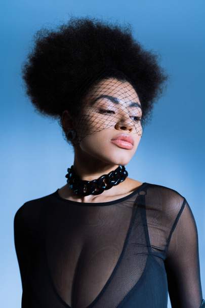 portrait of young african american woman in sexy outfit and black veil looking away while posing on blue  - Photo, Image