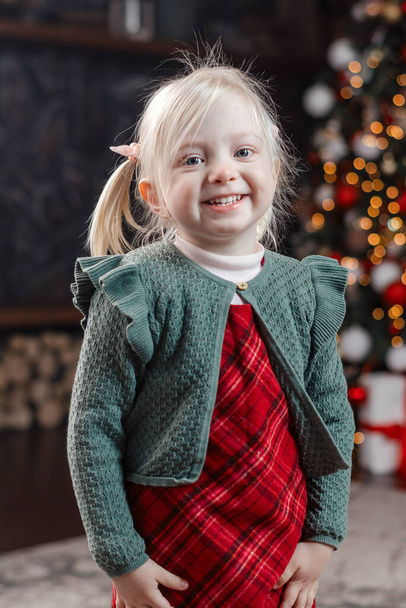 Portrait of little jolly blonde girl in festive new year clothes in traditional xmas decorated living room. Vertical frame. - Photo, Image