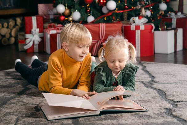 Fratellino biondo e sorella leggono un libro vicino all'albero di Natale. Fratelli e sorelle bambini la vigilia di Natale - Foto, immagini