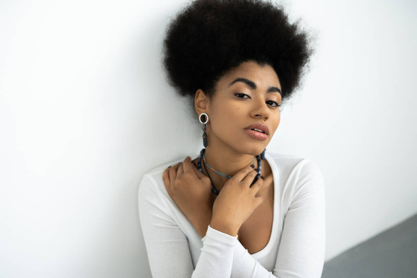 pretty african american woman with curly hair adjusting necklaces on white and grey - Foto, afbeelding