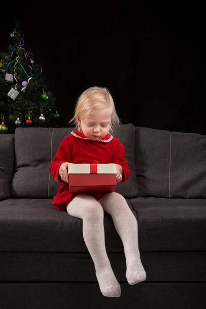 La bambina apre la confezione regalo seduta sul divano sullo sfondo di un albero di Natale. La vigilia di Natale. Regali di Capodanno. Telaio verticale. - Foto, immagini