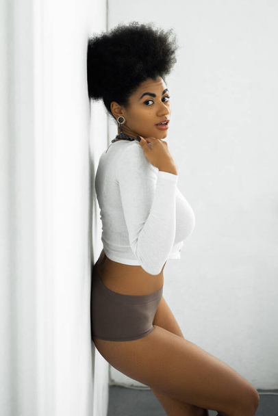mujer afroamericana bonita en camisa de manga larga y ropa interior de pie cerca de la pared blanca  - Foto, Imagen