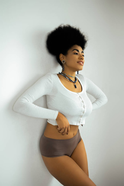 fröhliche afrikanisch-amerikanische Frau in langärmligem Hemd und Unterwäsche, die neben weißer Wand steht - Foto, Bild