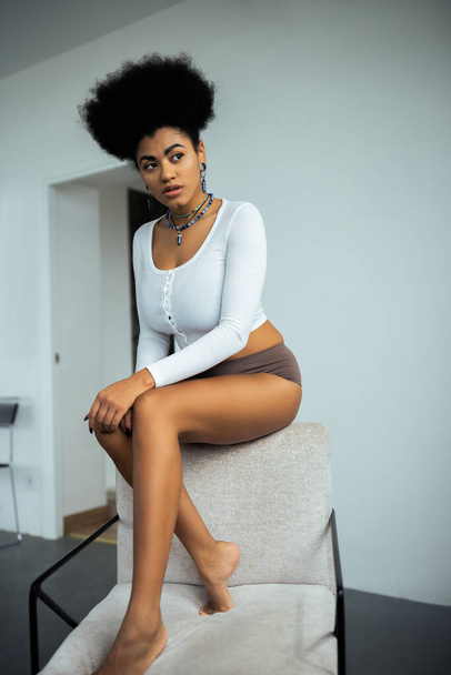 young african american woman in long sleeve shirt and panties looking away while sitting on armchair near white wall  - 写真・画像