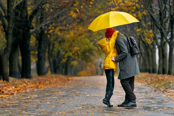 Zoon knuffelt zijn moeder lopend in het herfstpark in de regen met grote gele paraplu. Regendag. - Foto, afbeelding