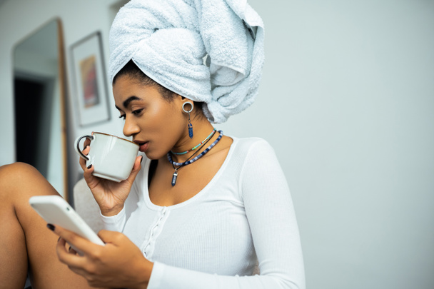 jolie femme américaine africaine avec serviette sur la tête en utilisant un smartphone et en buvant du café  - Photo, image