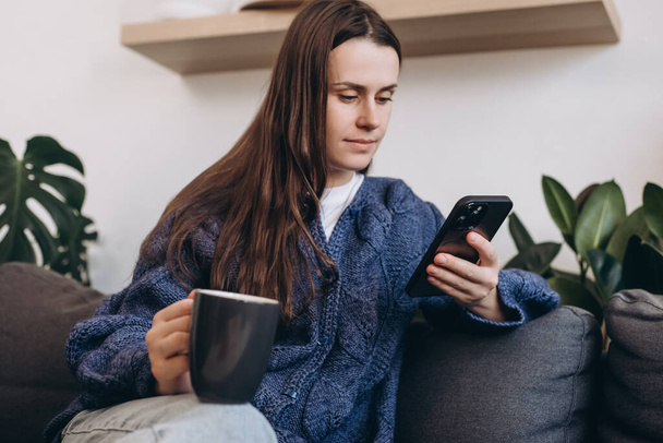 Selective focus of calm young woman sitting on couch holding cup and using smartphone. Beautiful brunette lady surfing internet, chatting, blogging. Close up girl hold phone and looking at screen - Zdjęcie, obraz