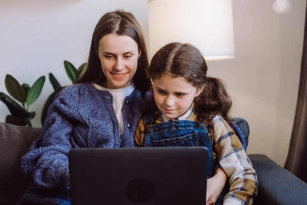 Selective focus of positive young mother with pretty little daughter using laptop, happy mum and cute girl child sitting on sofa in living room at home, watching cartoons online or play video game - Photo, Image