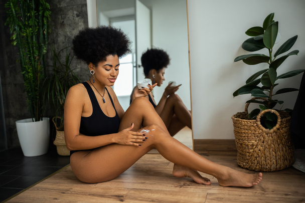 African american woman applying cosmetic cream on knee near mirror and plants in bathroom  - Fotografie, Obrázek