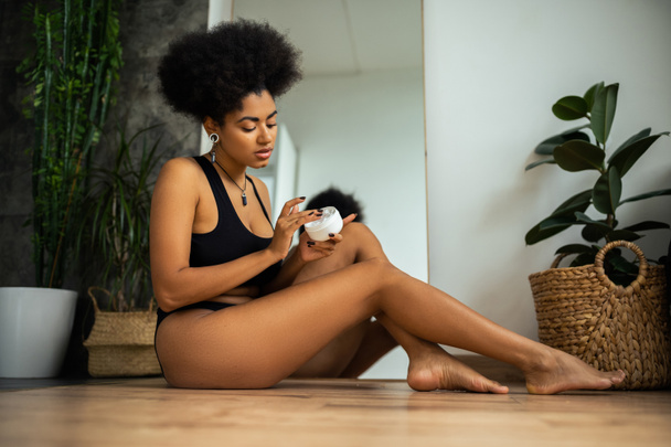 Jeune femme afro-américaine tenant de la crème cosmétique tout en étant assis sur le sol dans la salle de bain  - Photo, image