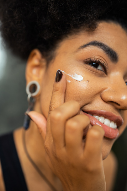 Cropped view of smiling african american woman applying cream on cheek at home  - Photo, Image