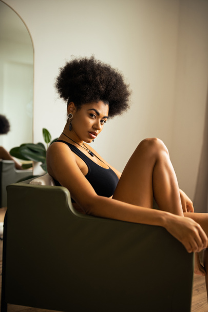 rizado africano americano mujer en negro crop top mirando cámara mientras sentado en sillón en casa  - Foto, imagen