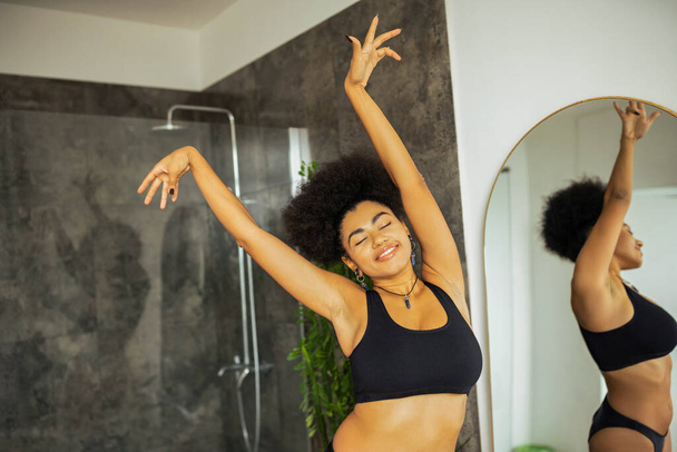 Femme afro-américaine gaie en sous-vêtements debout près du miroir et cabine de douche dans la salle de bain  - Photo, image