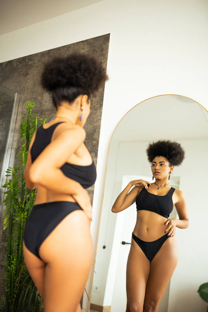Young african american woman touching body while looking at mirror in bathroom  - 写真・画像