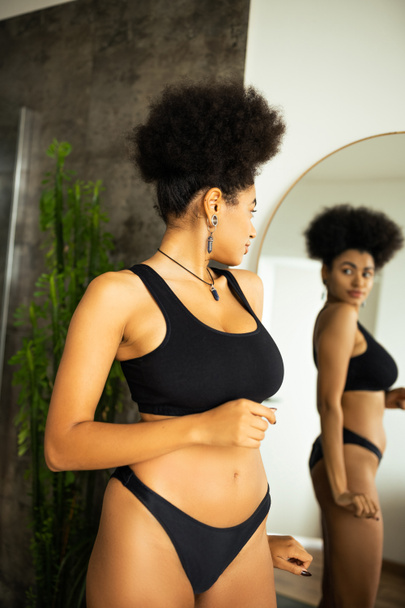 Side view of curly african american woman in underwear looking at blurred mirror at home  - Foto, Bild