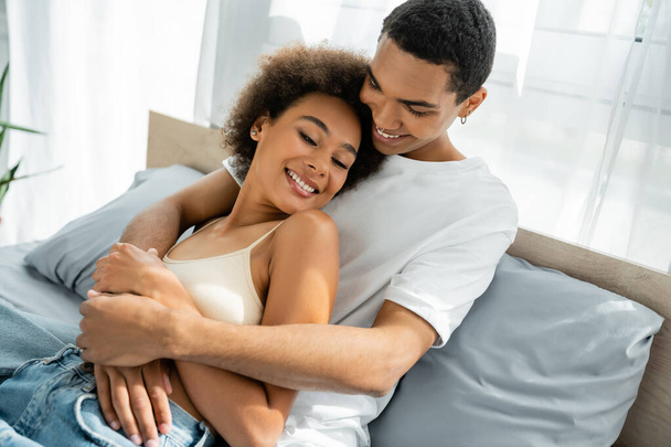 smiling african american man in white t-shirt embracing happy young girlfriend on bed at home - Fotó, kép