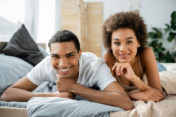 pierced african american man with young curly girlfriend lying on bed and smiling at camera - Фото, зображення