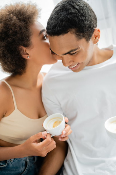 young african american woman with coffee cup telling secret to smiling boyfriend at home - Фото, изображение