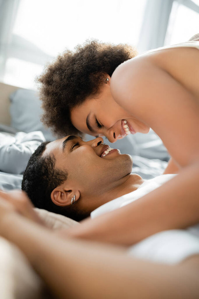 alegre africana americana mujer sonriendo sobre feliz novio en cama - Foto, Imagen