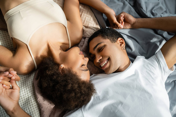 вид сверху на счастливую африканскую пару, держащуюся за руки, лежащую на кровати с закрытыми глазами - Фото, изображение