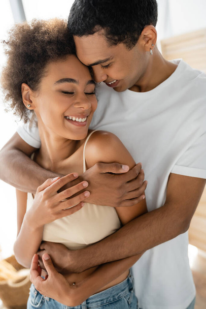 curly african american woman smiling with closed eyes near boyfriend hugging her at home - Photo, Image