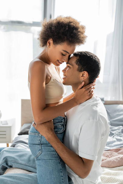 side view of african american woman in jeans and crop top hugging smiling boyfriend at home - Photo, Image