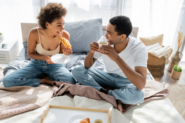 cheerful african american couple drinking coffee with croissant during breakfast in bedroom - Valokuva, kuva