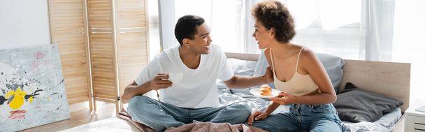 feliz pareja afroamericana con café y croissant hablando durante el desayuno en la cama, pancarta - Foto, Imagen