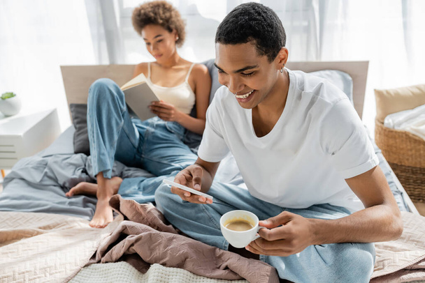 boldog afro-amerikai férfi kávéscsésze okostelefon közelében elmosódott barátnő olvasás könyv az ágyban  - Fotó, kép